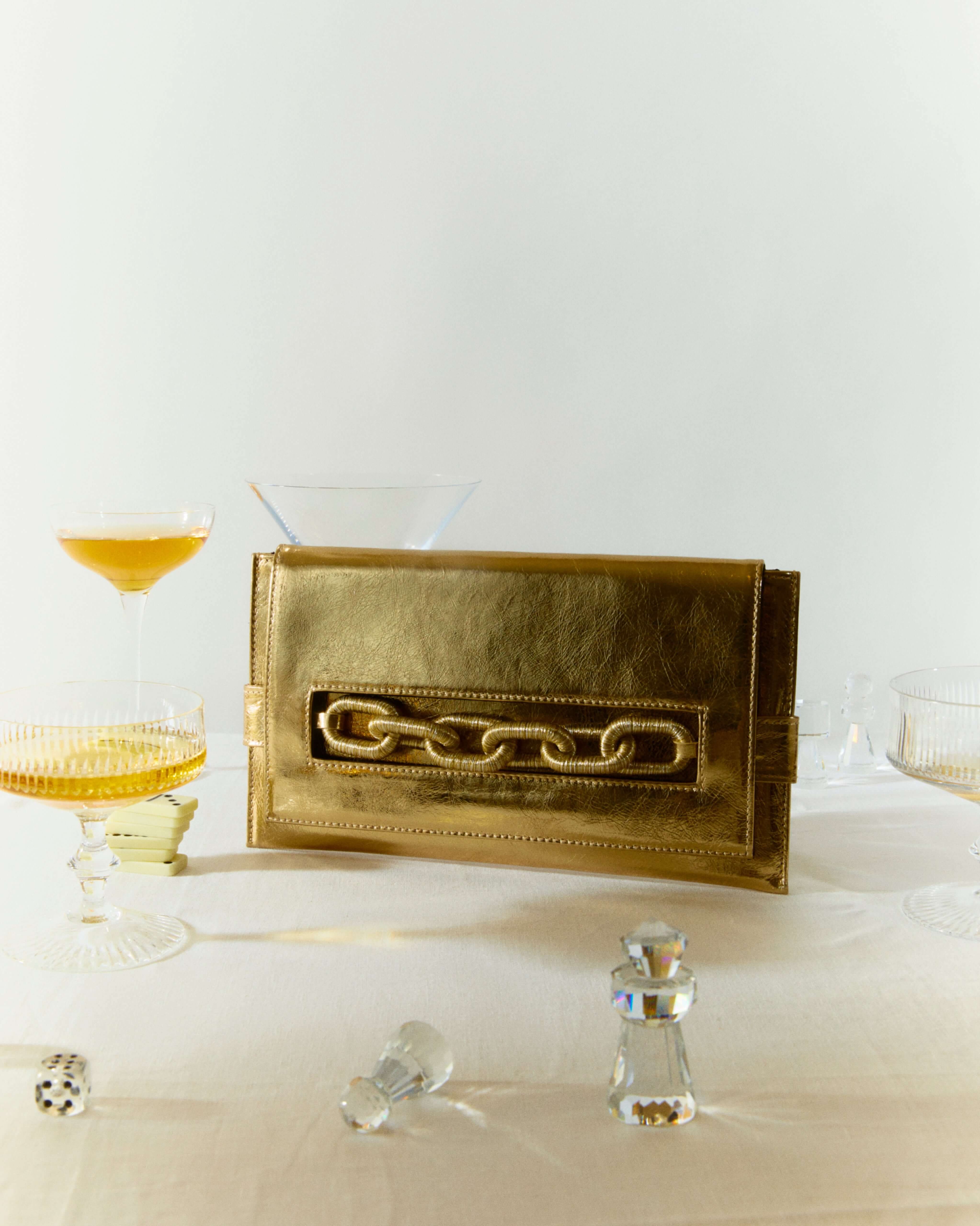 Catena Gold Envelope Clutch in Italian leather with chain embroidery, styled elegantly on a table with drinks.