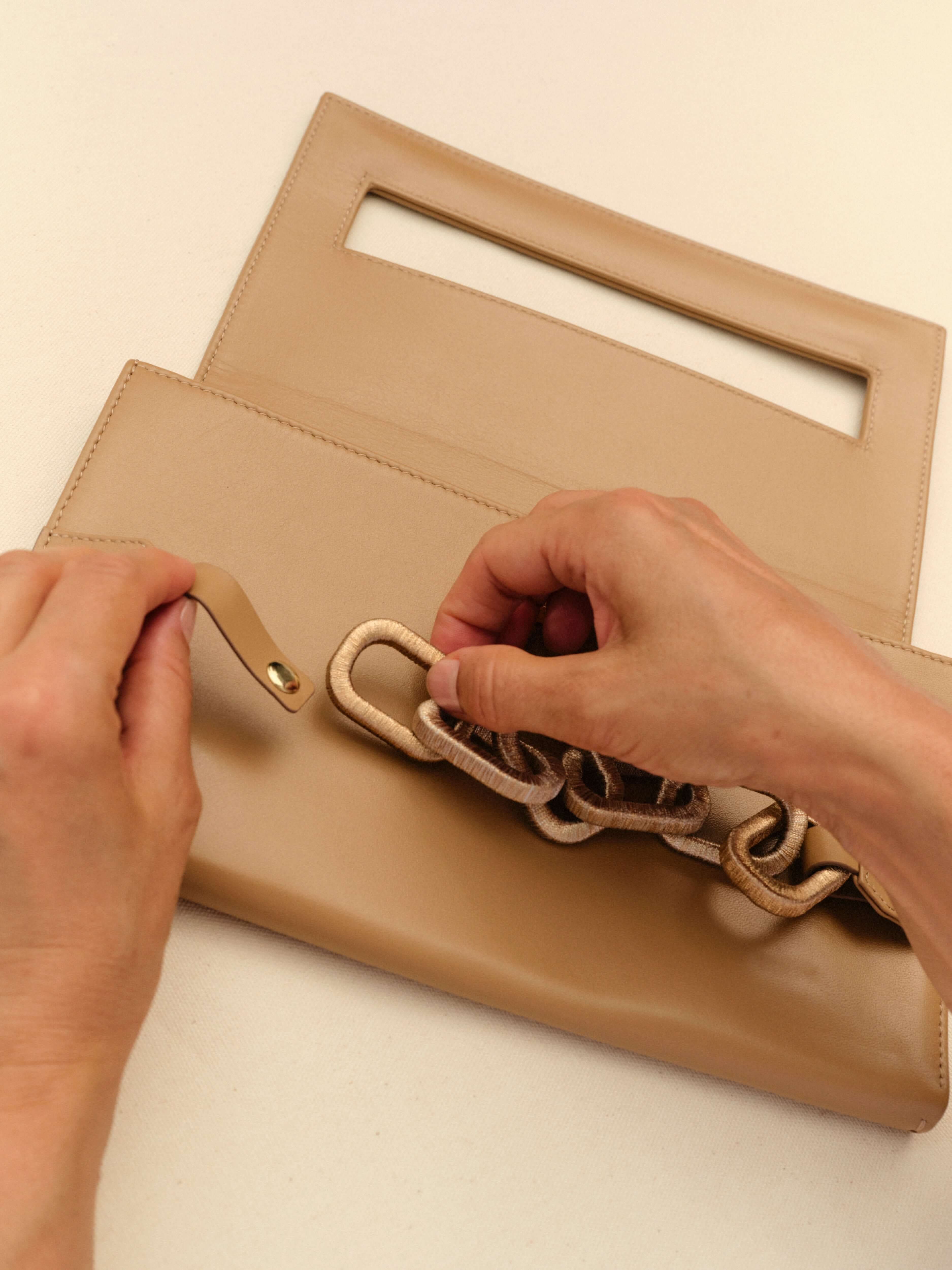 Hands attaching a stylish gold and silver chain to a sleek beige handbag, showcasing modern fashion design.