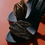 Close-up of elegant black high-heeled sandals with intricate leaf details on a vibrant orange background.