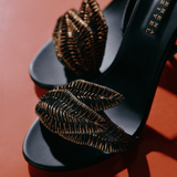 Close-up of elegant black high-heeled sandals with intricate leaf details on a vibrant orange background.