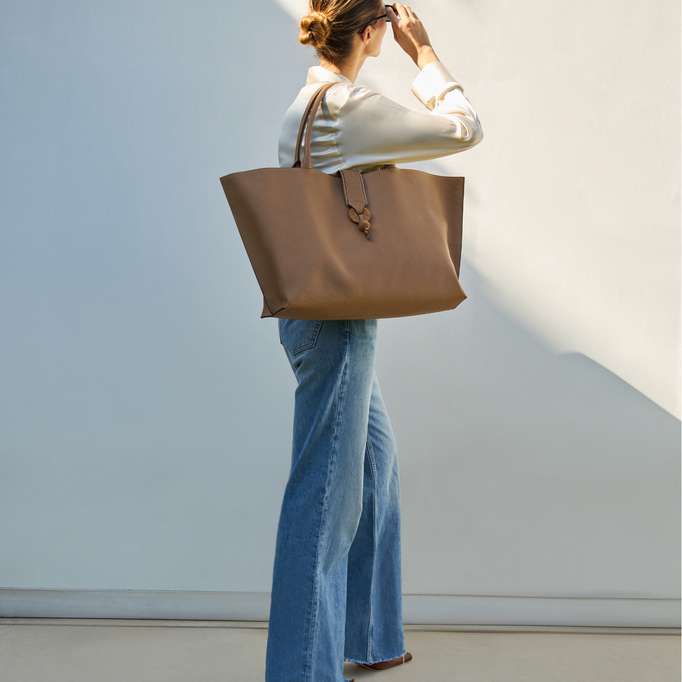 A model is carrying the cappuccino leather tote bag on her shoulder, wearing a white shirt and blue jeans with light brown shoes against a light wall background.