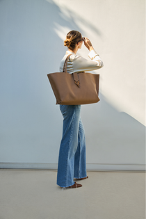 A model is carrying the cappuccino leather tote bag on her shoulder, wearing a white shirt and blue jeans with light brown shoes against a light wall background.