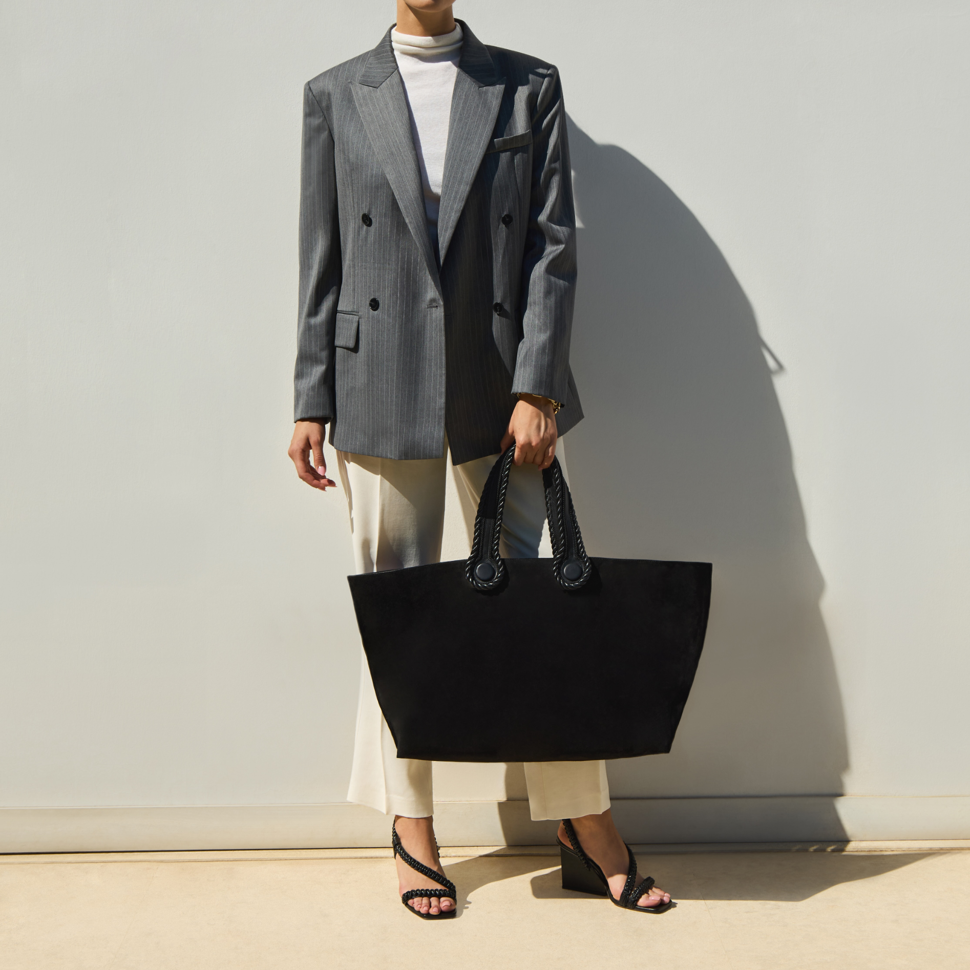 A model holding a black suede large tote bag and wearing a gray blazer,  white pants with black sandals against a plain background.