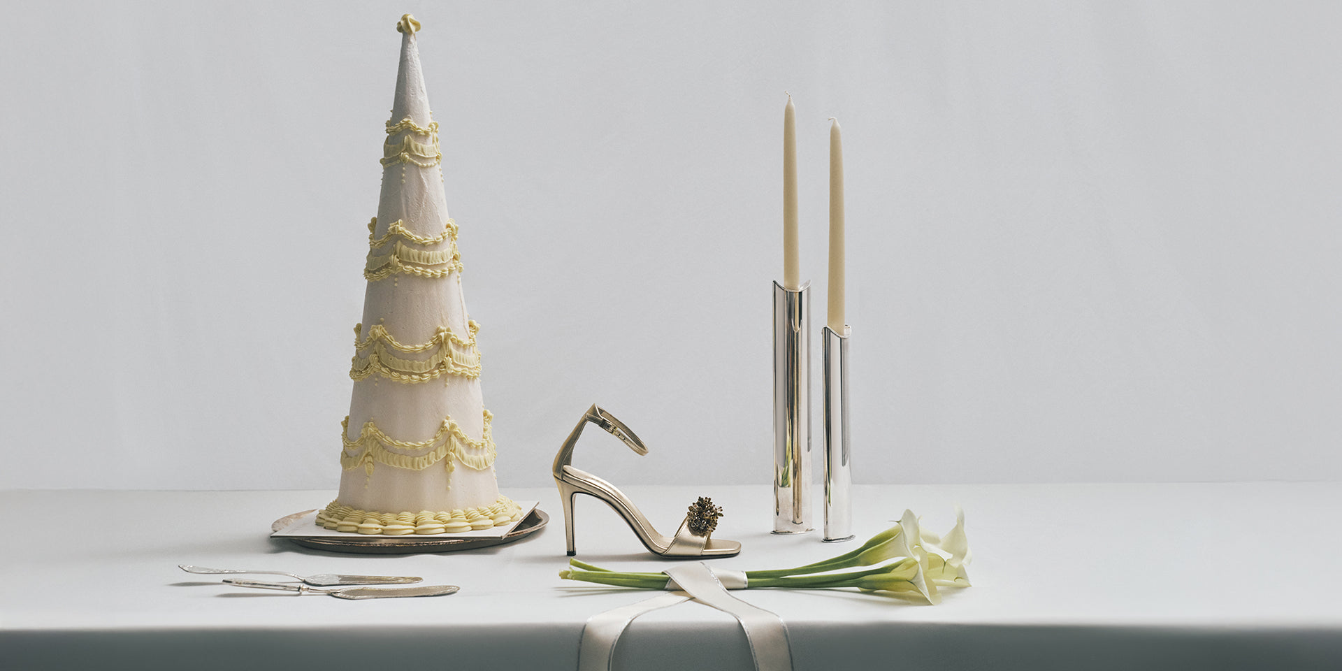 Decorative cake, gold high heel shoe, silver candle holder, and white flowers on a light gray surface.