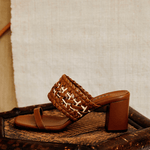 Almeria Camello Block-Heel Strap Sandal, crafted from supple camello leather, displayed on a rustic wooden chair.
