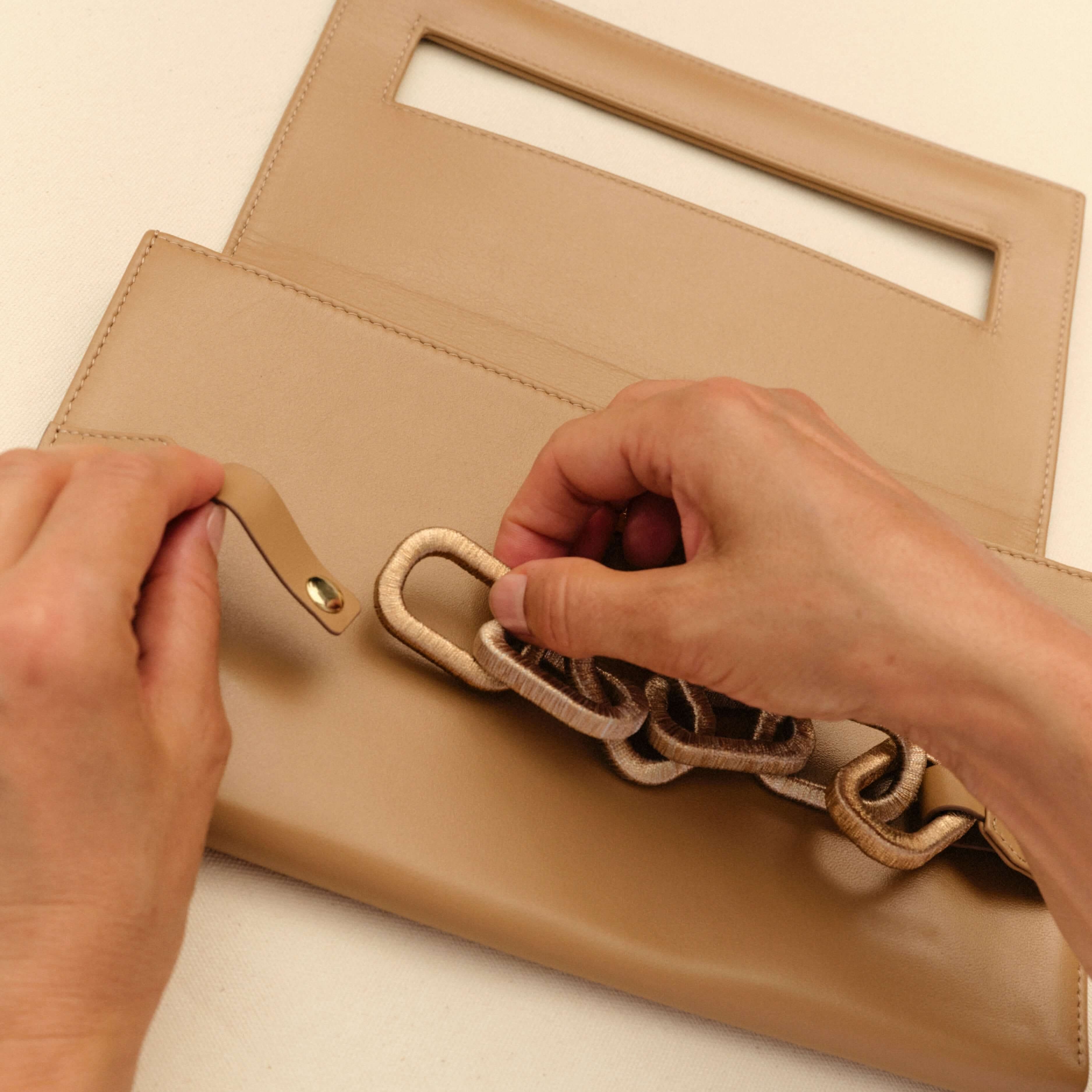Person attaching a chic blush chain to a stylish beige handbag, showcasing modern accessory design and craftsmanship.