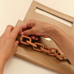 Hands attaching a Catena Raffia Chain to a stylish beige handbag, showcasing elegant design and craftsmanship.