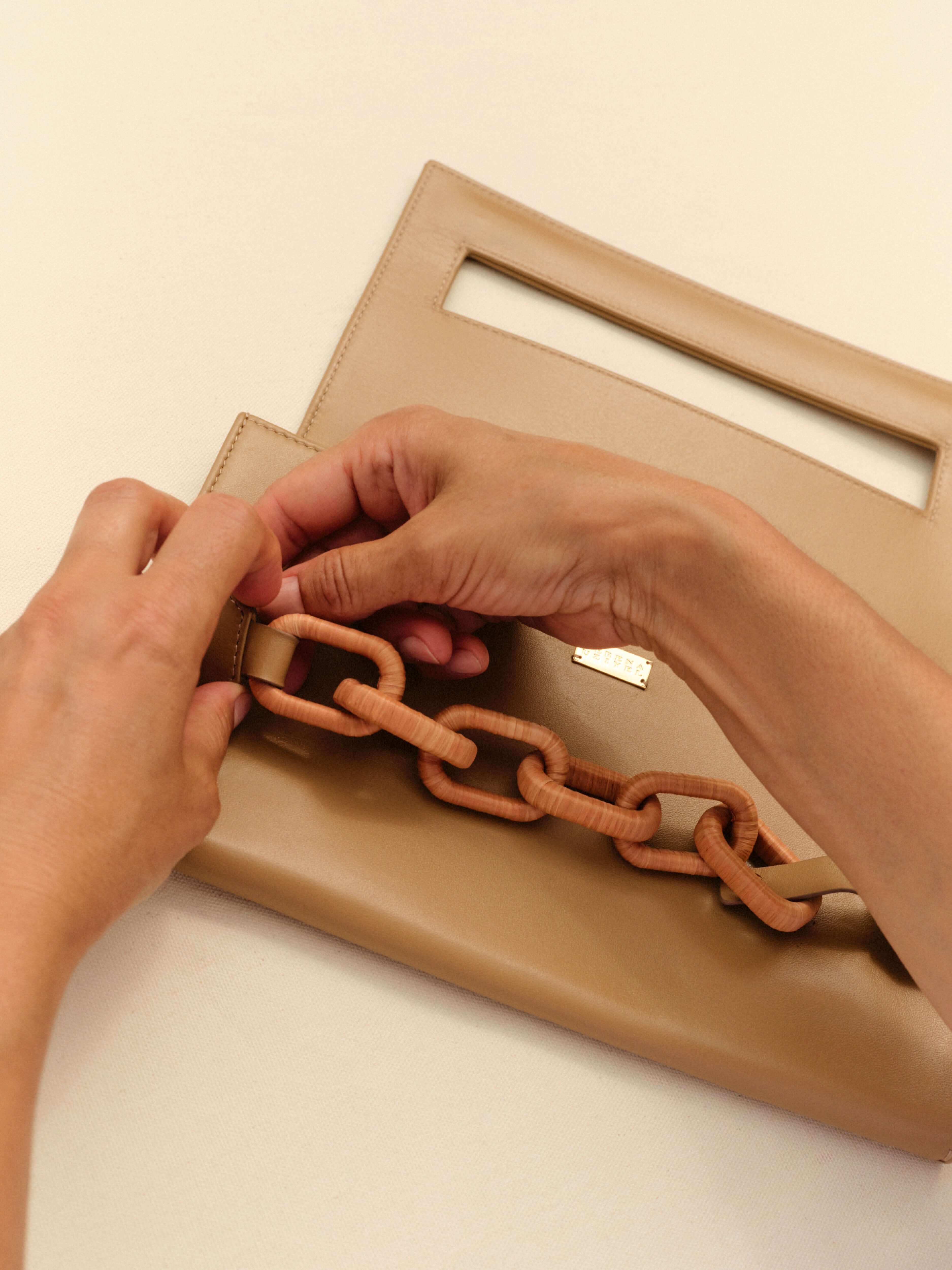 Hands attaching a Catena Raffia Chain to a stylish beige handbag, showcasing elegant design and craftsmanship.