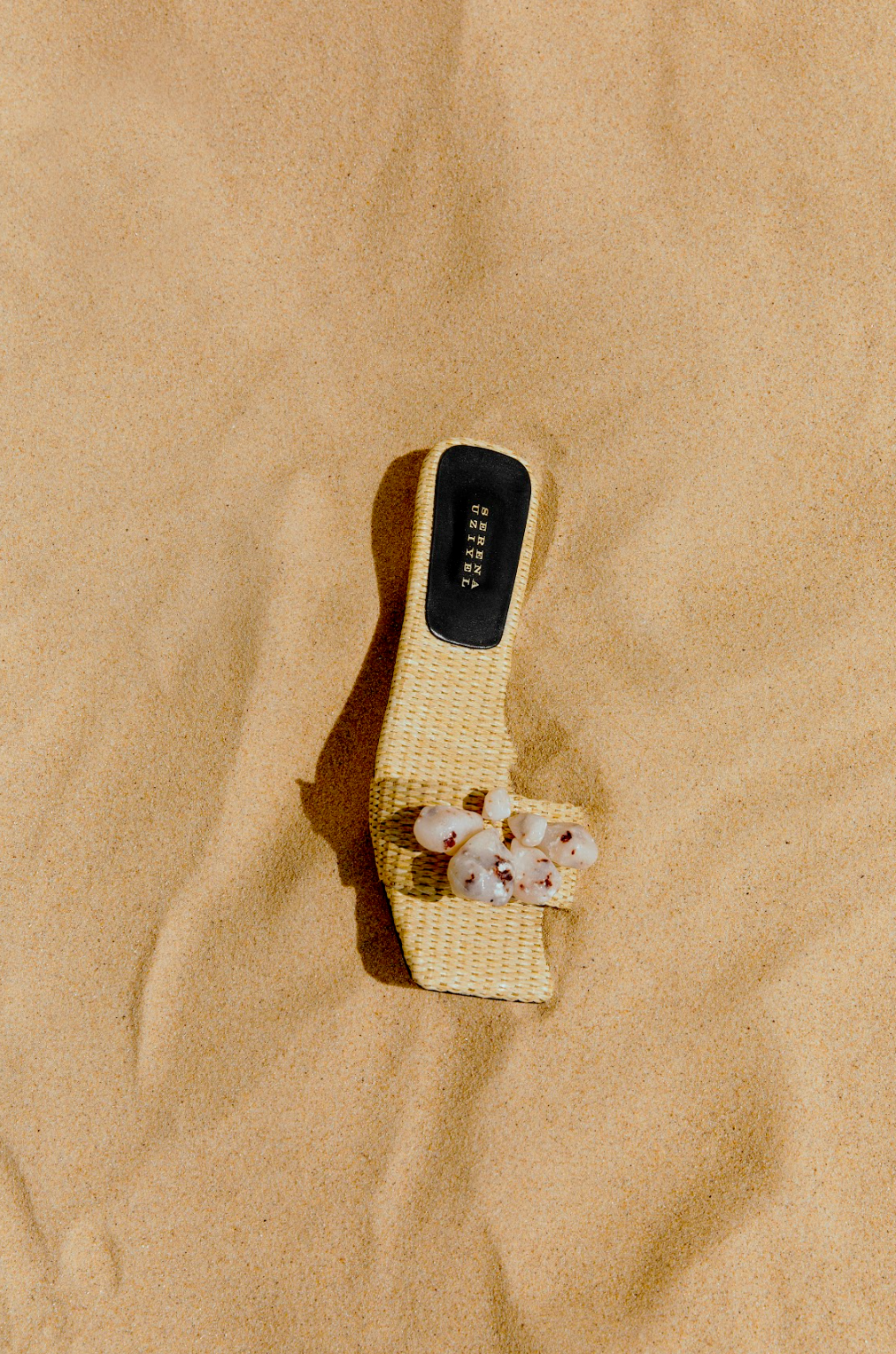 Taurea Natural Flat Sandal in sand colored raffia with stone embellishments on beach sand.
