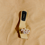 Taurea Natural Flat Sandal in sand colored raffia with stone embellishments on beach sand.